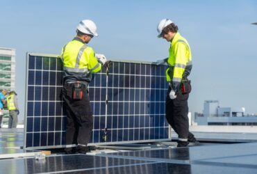 Two technicians in high-visibility jackets and helmets carefully install a solar panel on a rooftop with urban buildings in the background.