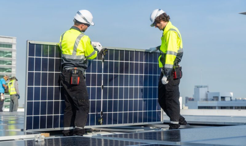 Two technicians in high-visibility jackets and helmets carefully install a solar panel on a rooftop with urban buildings in the background.